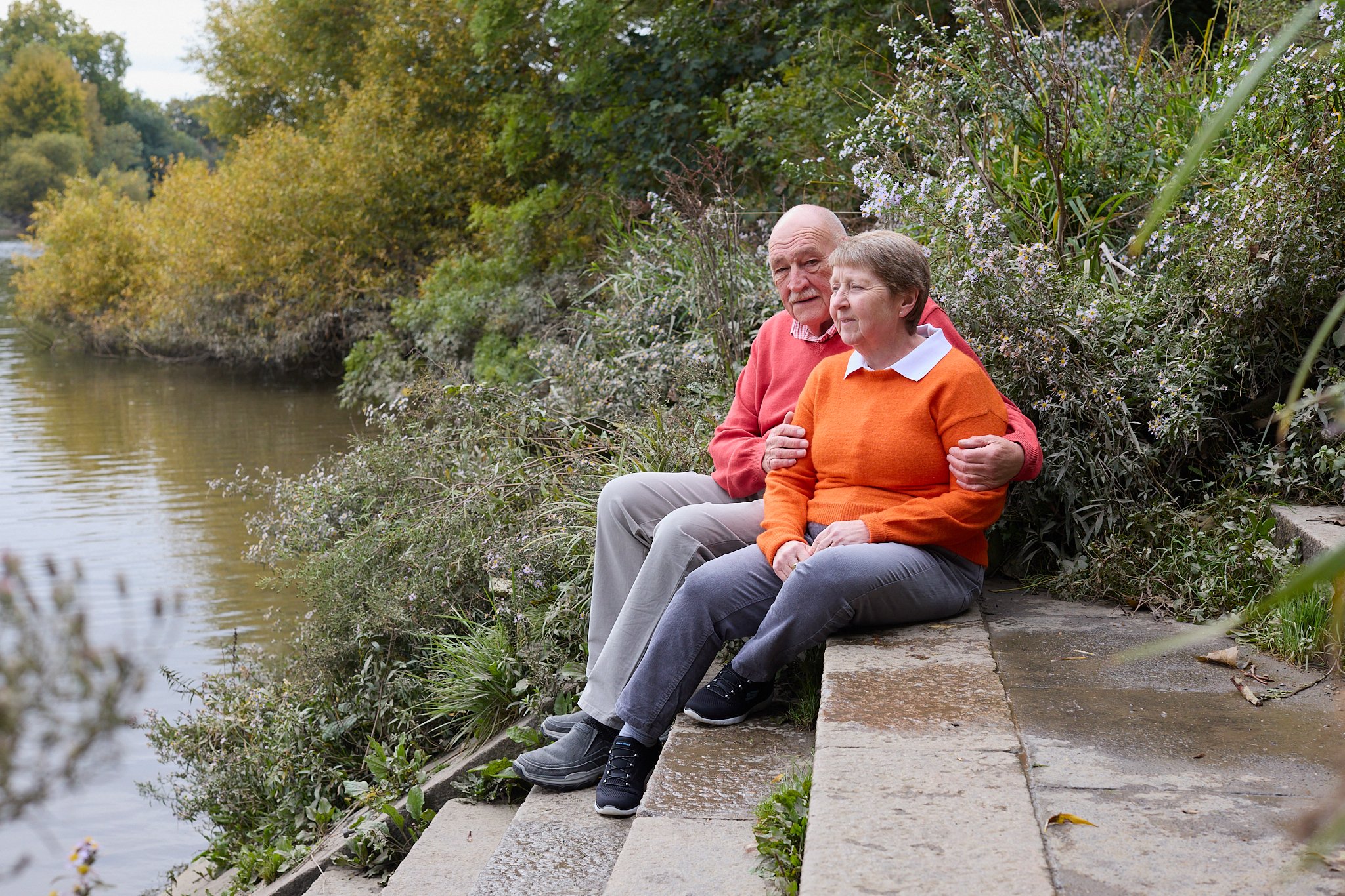An older couple sits close together on a concrete riverside path, the woman in orange and man in red, surrounded by greenery; calm, affectionate mood.