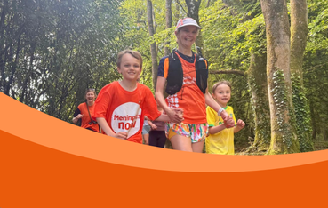 White woman in cap and running vest holds hands with two little boys with a background of trees