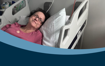 white woman with dark hair who's wearing glasses and a pink top lies in a hospital bed with her eyes open