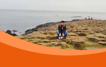 A young man crouches on a rocky coastal shore, with calm grey sea and distant walkers behind him; an orange graphic curves across the foreground.