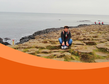 A young man crouches on a rocky coastal shore, with calm grey sea and distant walkers behind him; an orange graphic curves across the foreground.
