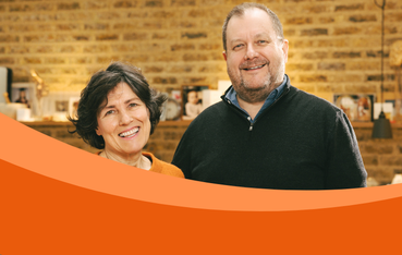 White couple stand in front of brick wall. Woman on left with dark hair smiling, man on right with dark navy jumper