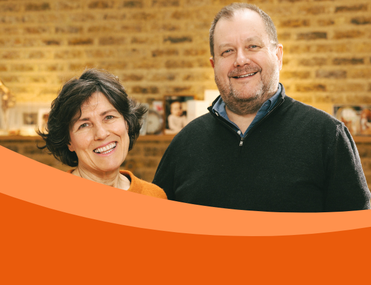 White couple stand in front of brick wall. Woman on left with dark hair smiling, man on right with dark navy jumper