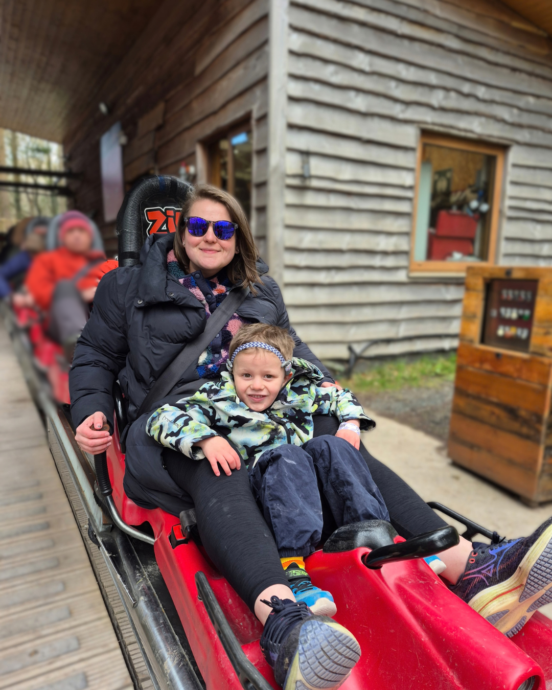 A smiling woman in sunglasses rides a red alpine coaster with a happy toddler strapped in front; cabins and trackside buildings create a cheerful outdoor setting.