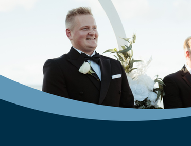 White man with blonde hair wearing a smart suit smiles, looking just out of frame and surrounded by flowers