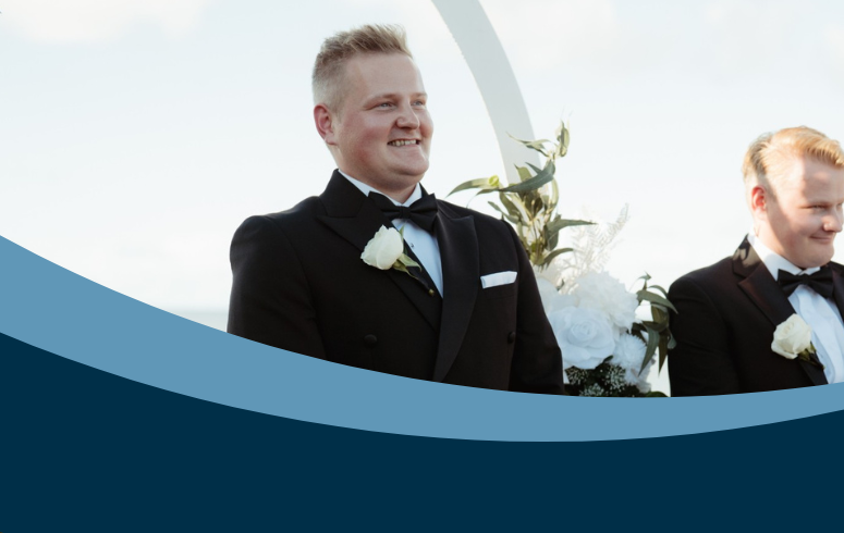 White man with blonde hair wearing a smart suit smiles, looking just out of frame and surrounded by flowers