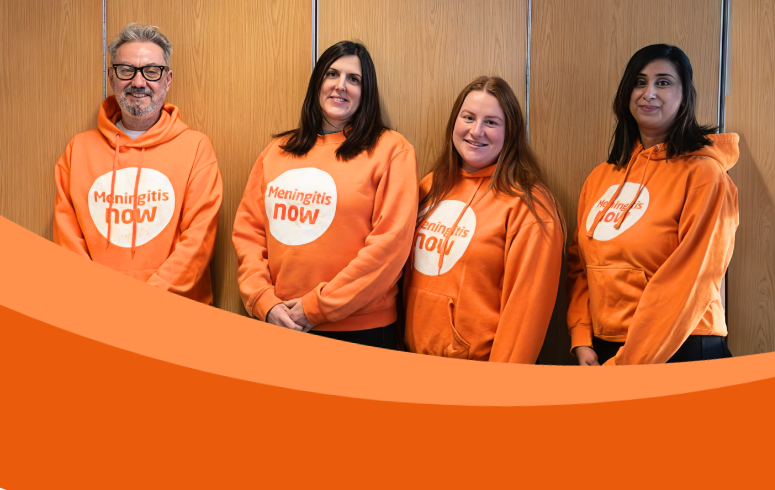 Four people stand in front of wooden wall wearing orange Meningitis Now hoodies