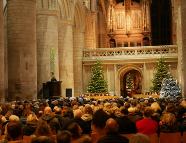 A large audience sits in a cathedral during a Christmas service, featuring a pipe organ and illuminated Christmas trees.