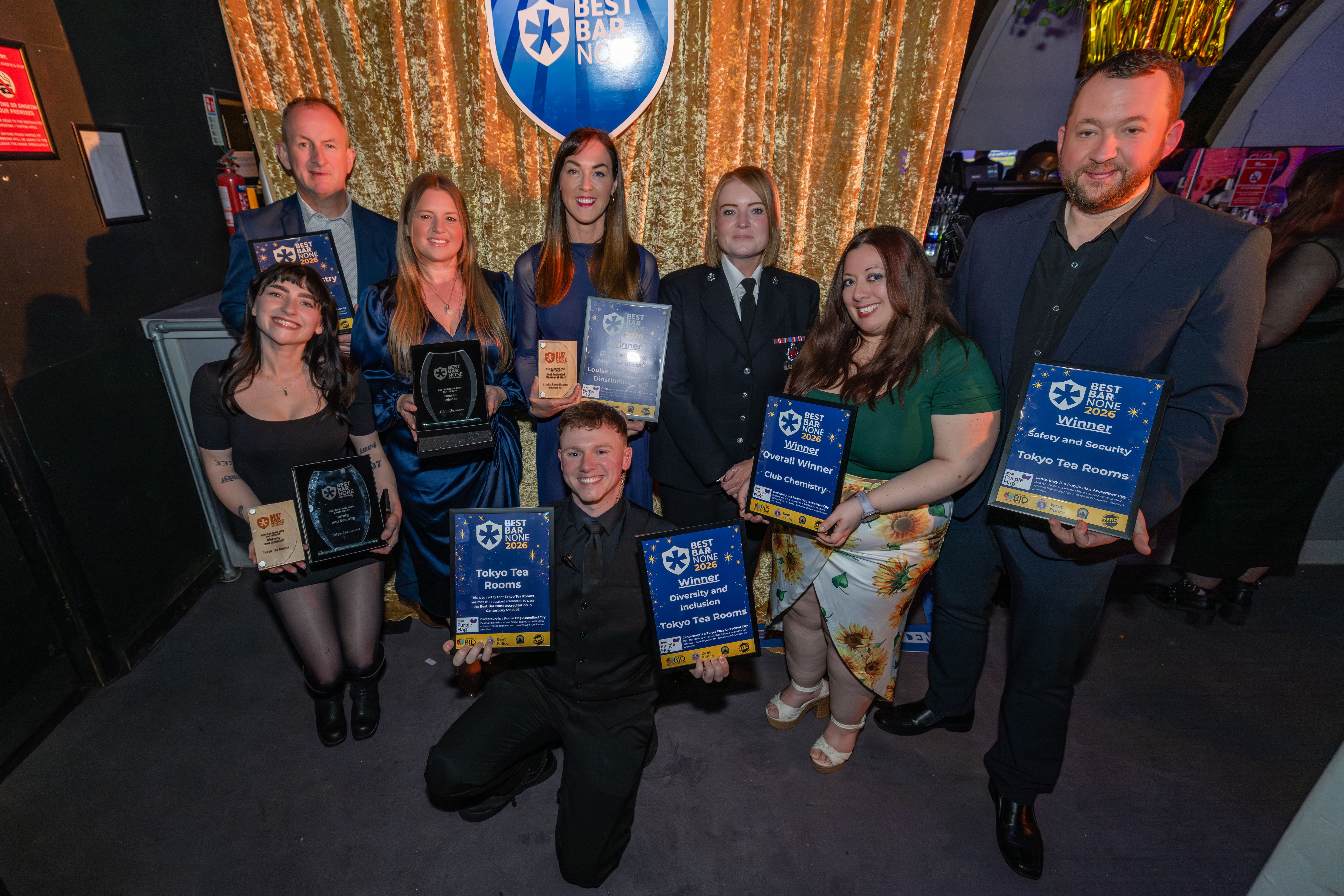 A group of eight people smiling and holding award plaques in front of a shiny gold curtain.