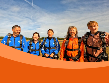 Five white people of different ages stand smiling at the camera - three are wearing blue skydiving suits and harnesses, and two are wearing orange