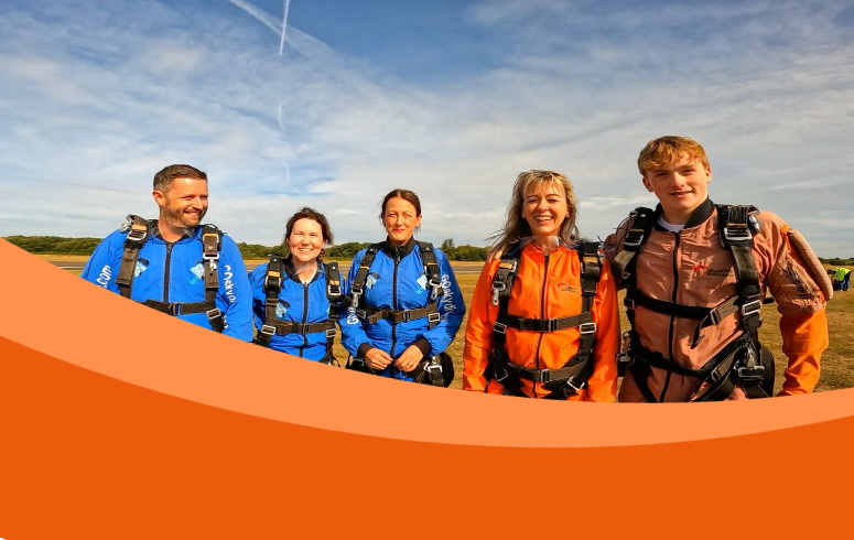 Five white people of different ages stand smiling at the camera - three are wearing blue skydiving suits and harnesses, and two are wearing orange