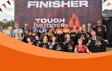 20 people covered in mud and wearing orange headbands and black t-shirts stand in front a board that reads 'Finisher: Tough Mudder'