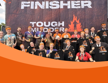 20 people covered in mud and wearing orange headbands and black t-shirts stand in front a board that reads 'Finisher: Tough Mudder'