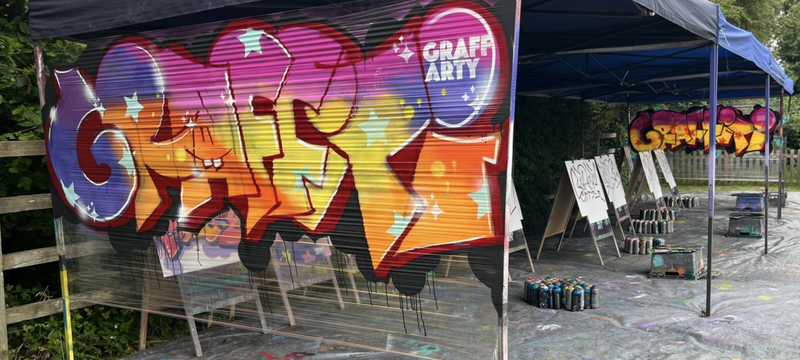Outdoor graffiti art booth with colorful spray-painted murals, paint cans, chairs, and splattered pavement under a canopy; vibrant creative mood.