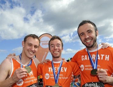 Three people dressed in orange meningitis now running tops showing their medals to camera after completing a race.