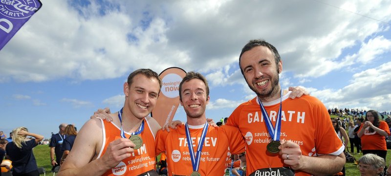 Three people dressed in orange meningitis now running tops showing their medals to camera after completing a race.