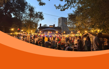 A lively outdoor festival scene with a crowd gathered in front of a stage adorned with colourful lights and flags. In the background, there is a historic stone church, partially visible through the trees. It's evening, and the sky is clear