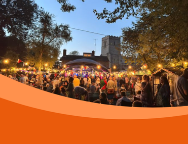 A lively outdoor festival scene with a crowd gathered in front of a stage adorned with colourful lights and flags. In the background, there is a historic stone church, partially visible through the trees. It's evening, and the sky is clear