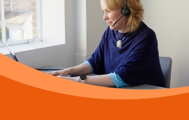 White woman with blonde hair looks away from camera to laptop screen. She is wearing a navy top and a headset.