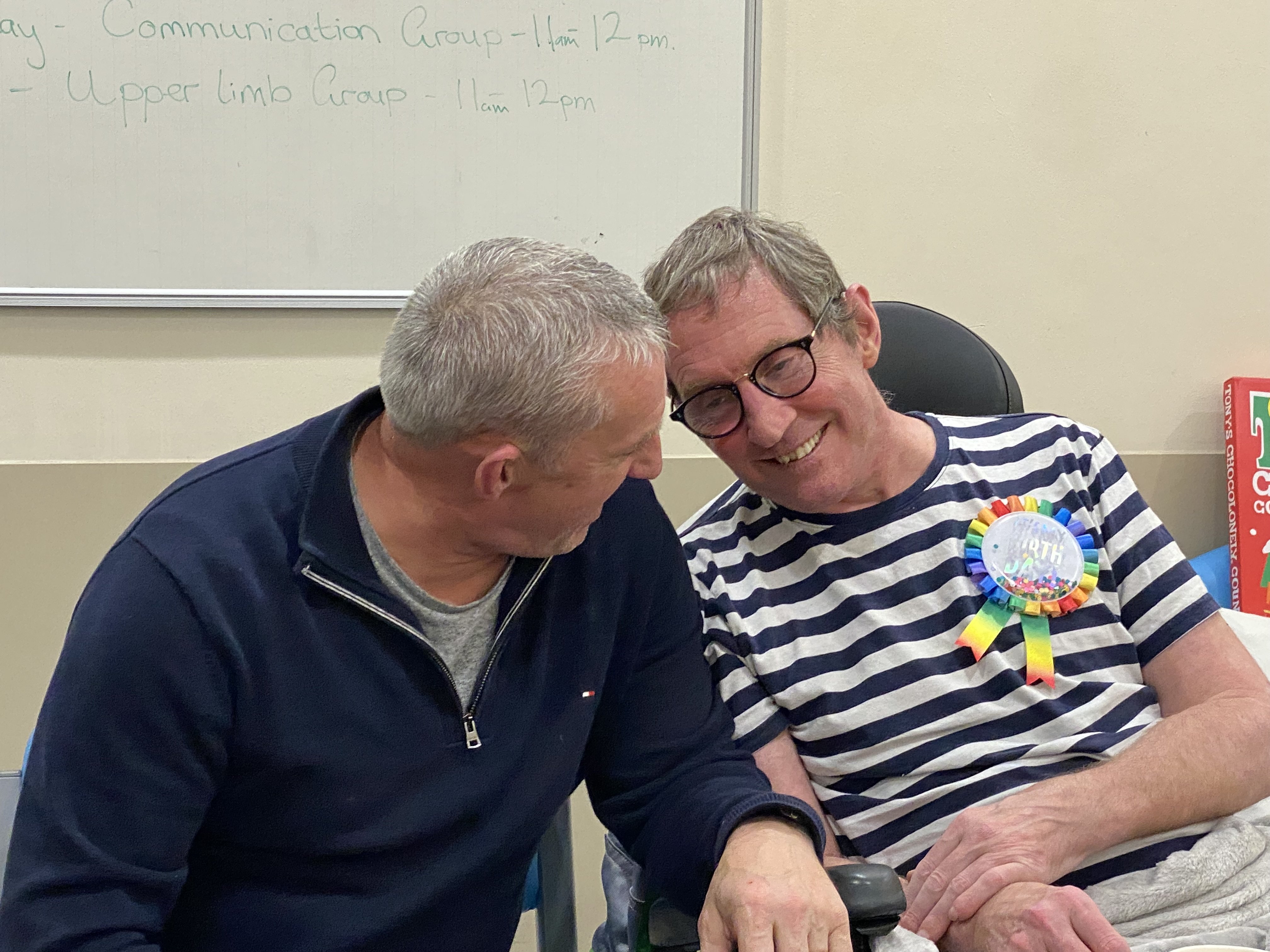 Two men sit close together in a hospital, smiling; one wears a striped shirt and rainbow rosette, while the other looks toward him warmly.