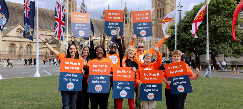 A group of people holding 'No Plan B for Men B' campaign signs outside of parliament buildings. The group are cheering with their hands up in the air.