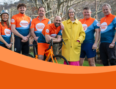 Group of seven white people in Meningitis Now sports vest smile at camera. CEO Tom Nutt is in the middle of the group on a bike.