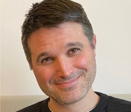 A man with stubble smiling to the camera wearing a dark grey sweater