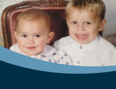 Two white children with blonde hair sit together on a wooden chair smiling at the camera