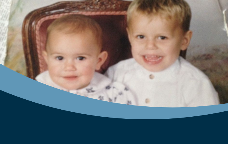 Two white children with blonde hair sit together on a wooden chair smiling at the camera