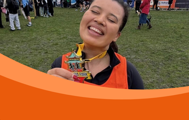 The image shows a smiling woman wearing an orange vest, holding up a medal with the words "Victoria Park" visible. She appears to be celebrating her accomplishment, standing on grass with event banners in the background.