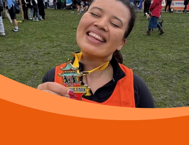 The image shows a smiling woman wearing an orange vest, holding up a medal with the words "Victoria Park" visible. She appears to be celebrating her accomplishment, standing on grass with event banners in the background.
