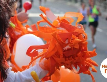 A supporter in an orange shirt holds bright orange pom-poms and balloons at a marathon, cheering for runners in the background. The atmosphere is energetic.