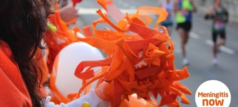 A supporter in an orange shirt holds bright orange pom-poms and balloons at a marathon, cheering for runners in the background. The atmosphere is energetic.