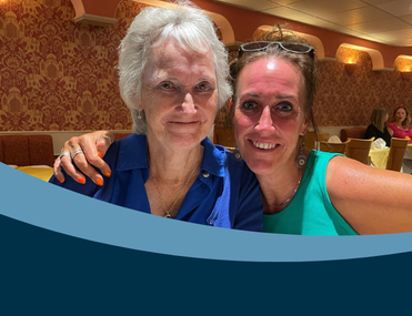 Two women smiling in a warmly lit restaurant, embracing each other. One wears a blue shirt, the other a green top. The atmosphere is cozy and cheerful.