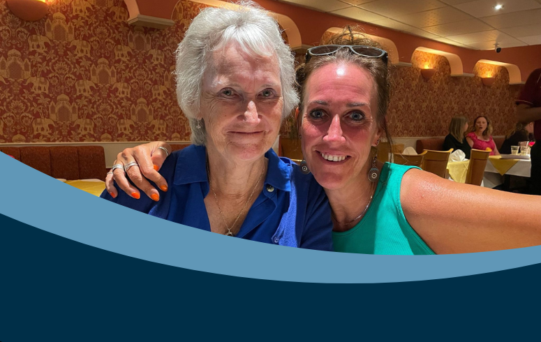 Two women smiling in a warmly lit restaurant, embracing each other. One wears a blue shirt, the other a green top. The atmosphere is cozy and cheerful.