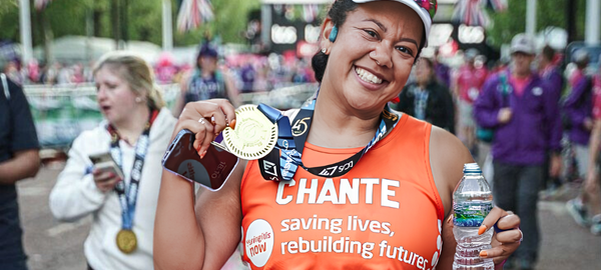 Smiling marathon finisher holds a medal and water bottle, wearing an orange "Chante" jersey. Other runners and Union Jack flags in the background.
