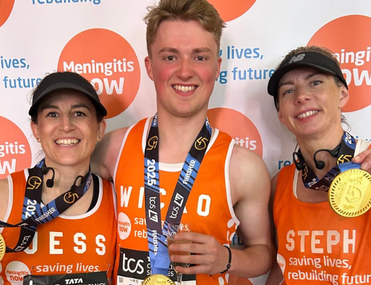 Three smiling runners wearing orange shirts and medals stand in front of a "Meningitis Now" backdrop with a joyful, celebratory tone.