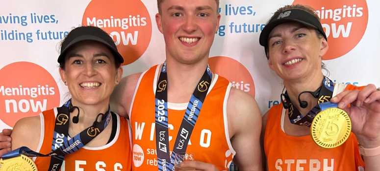 Three smiling runners wearing orange shirts and medals stand in front of a "Meningitis Now" backdrop with a joyful, celebratory tone.