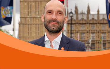 A man stands in a suit infront of a parliament building, with a Meninigitis Now badge. The photo has an orange sweeping border.