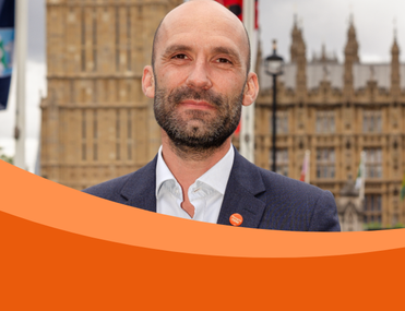 A man stands in a suit infront of a parliament building, with a Meninigitis Now badge. The photo has an orange sweeping border.