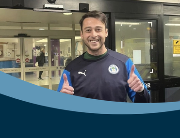 A smiling man in a sports jacket gives thumbs up outside a building with glass doors.