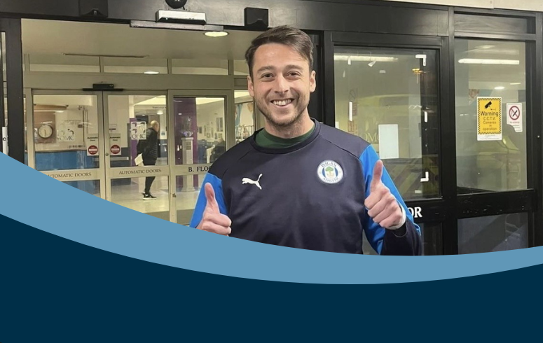 A smiling man in a sports jacket gives thumbs up outside a building with glass doors.