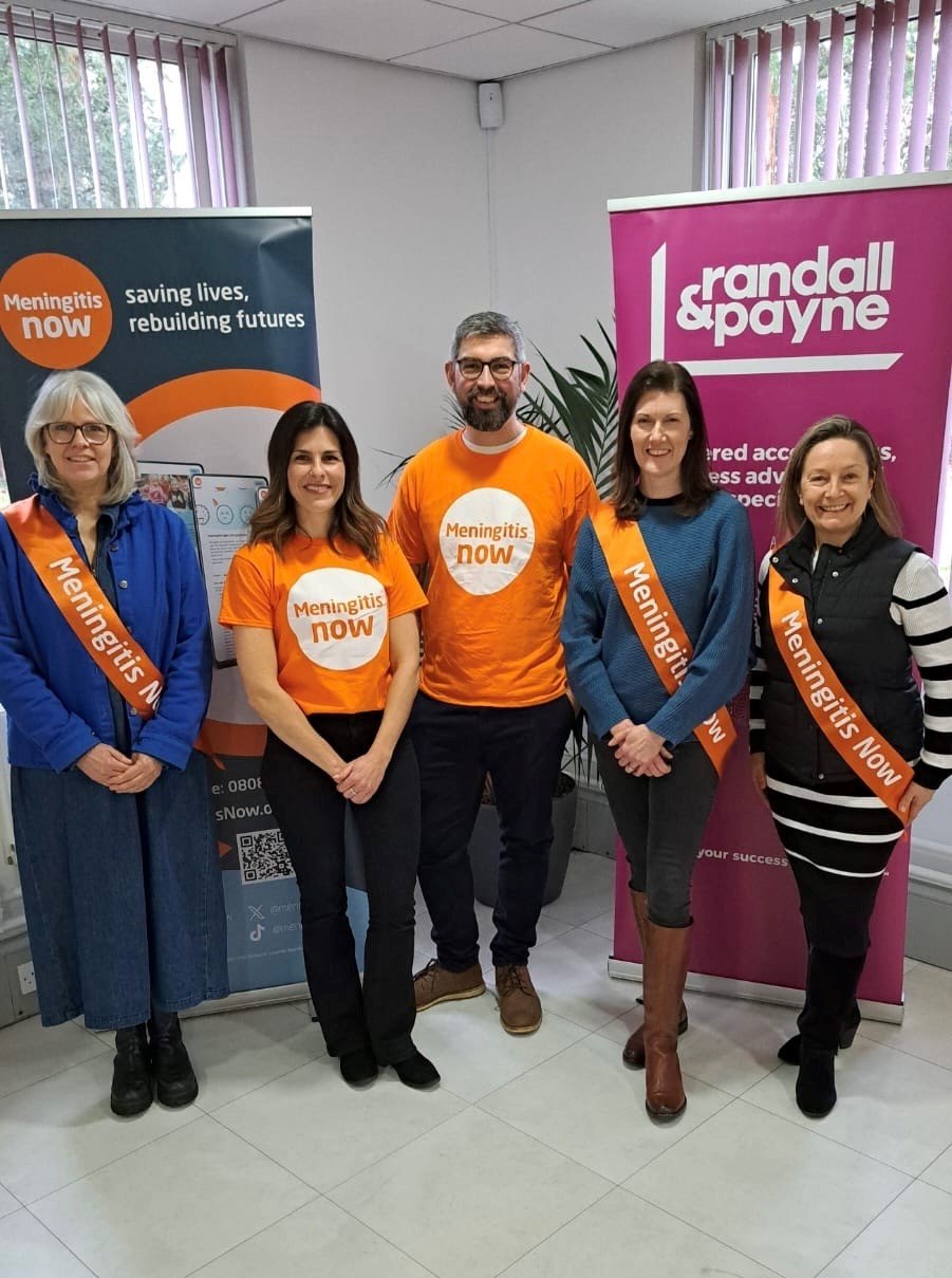 Five people stand smiling in front of banners for "Meningitis Now" and "randall & payne," wearing branded sashes and T-shirts, conveying a positive and collaborative tone.