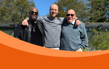 Three men standing with arms around each other - from left to right, a Black man wearing sunglasses, black clothes and a silver cross necklace, a taller white man wearing a marl grey t-shirt and an older white man with moustache