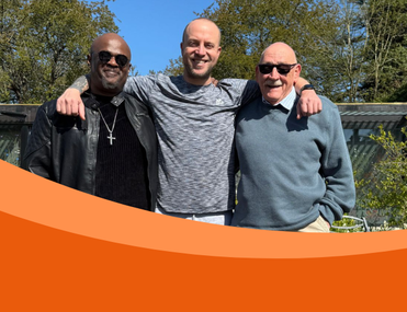 Three men standing with arms around each other - from left to right, a Black man wearing sunglasses, black clothes and a silver cross necklace, a taller white man wearing a marl grey t-shirt and an older white man with moustache