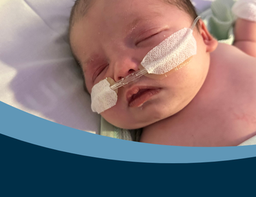 A newborn resting on a hospital bed, swaddled in a light green blanket. The infant is wearing nasal cannulas and has medical tape on the face, with mittens on the hands. A transparent dummy lies nearby.