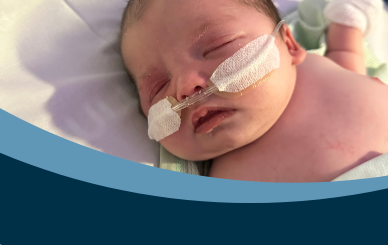 A newborn resting on a hospital bed, swaddled in a light green blanket. The infant is wearing nasal cannulas and has medical tape on the face, with mittens on the hands. A transparent dummy lies nearby.