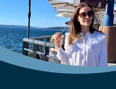 white woman with brown hair blowing in the wind, wearing a white blouse standing in front of outdoor seating and the sea beyond