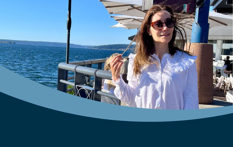 white woman with brown hair blowing in the wind, wearing a white blouse standing in front of outdoor seating and the sea beyond