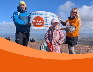 Young girl in pink hat and coat stands in front of a banner held by her parents at the top of a mountain
